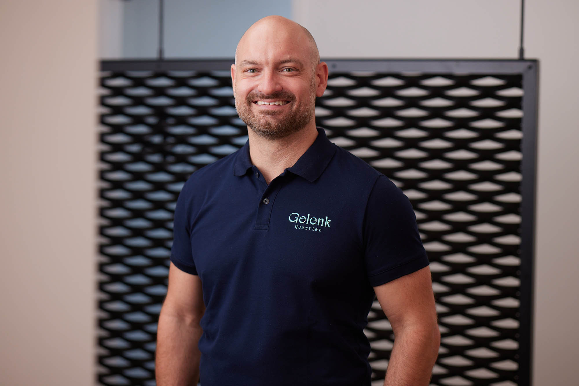 Dr. Zimmerer lächelt vor einer Wand stehend in die Kamera. Er trägt ein dunkelblaues Poloshirt mit dem Logo „Gelenkquartier“.