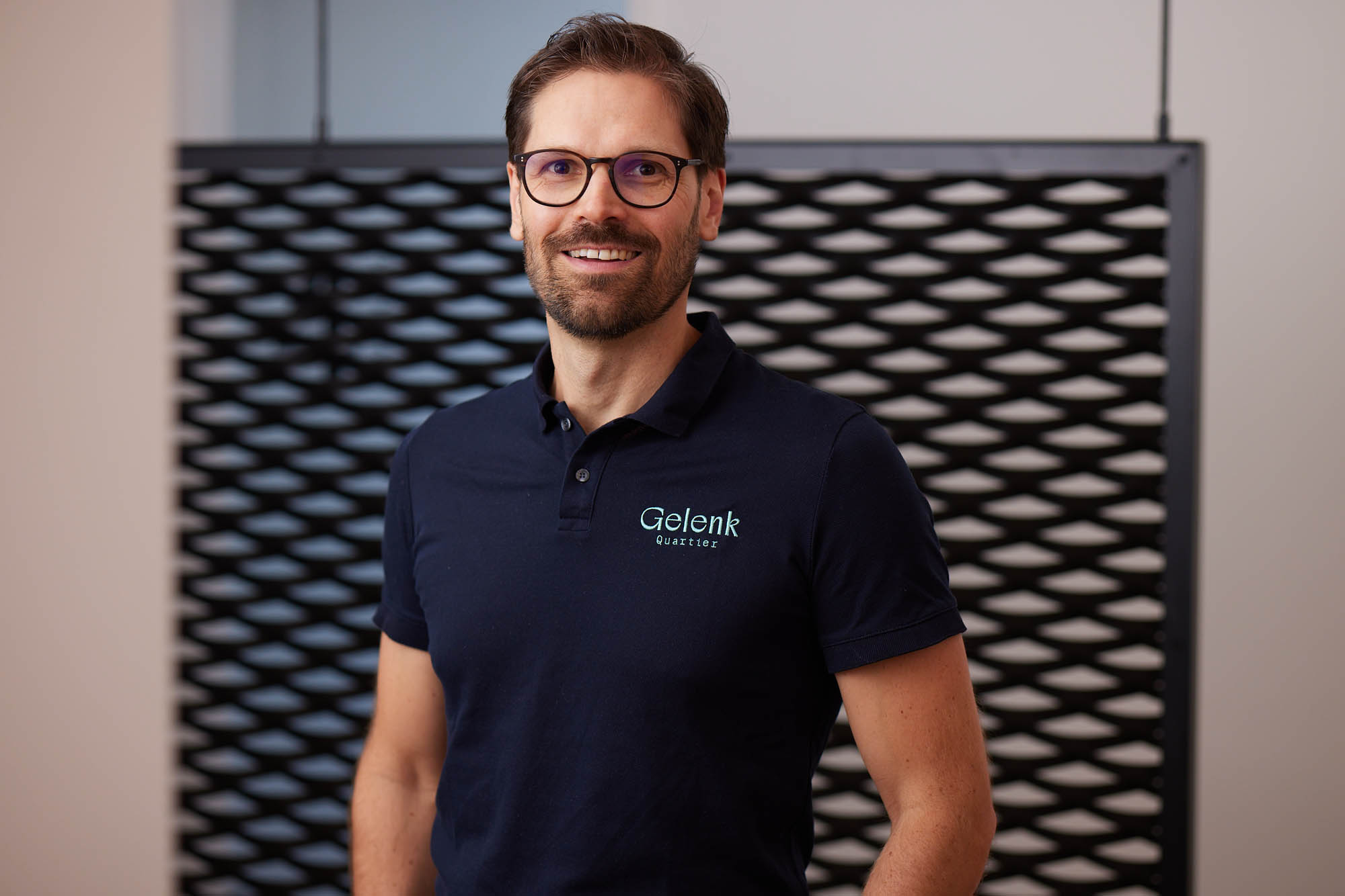 Dr. Nietschke lächelt vor einer Wand stehend in die Kamera. Er trägt ein dunkelblaues Poloshirt mit dem Logo „Gelenkquartier“.