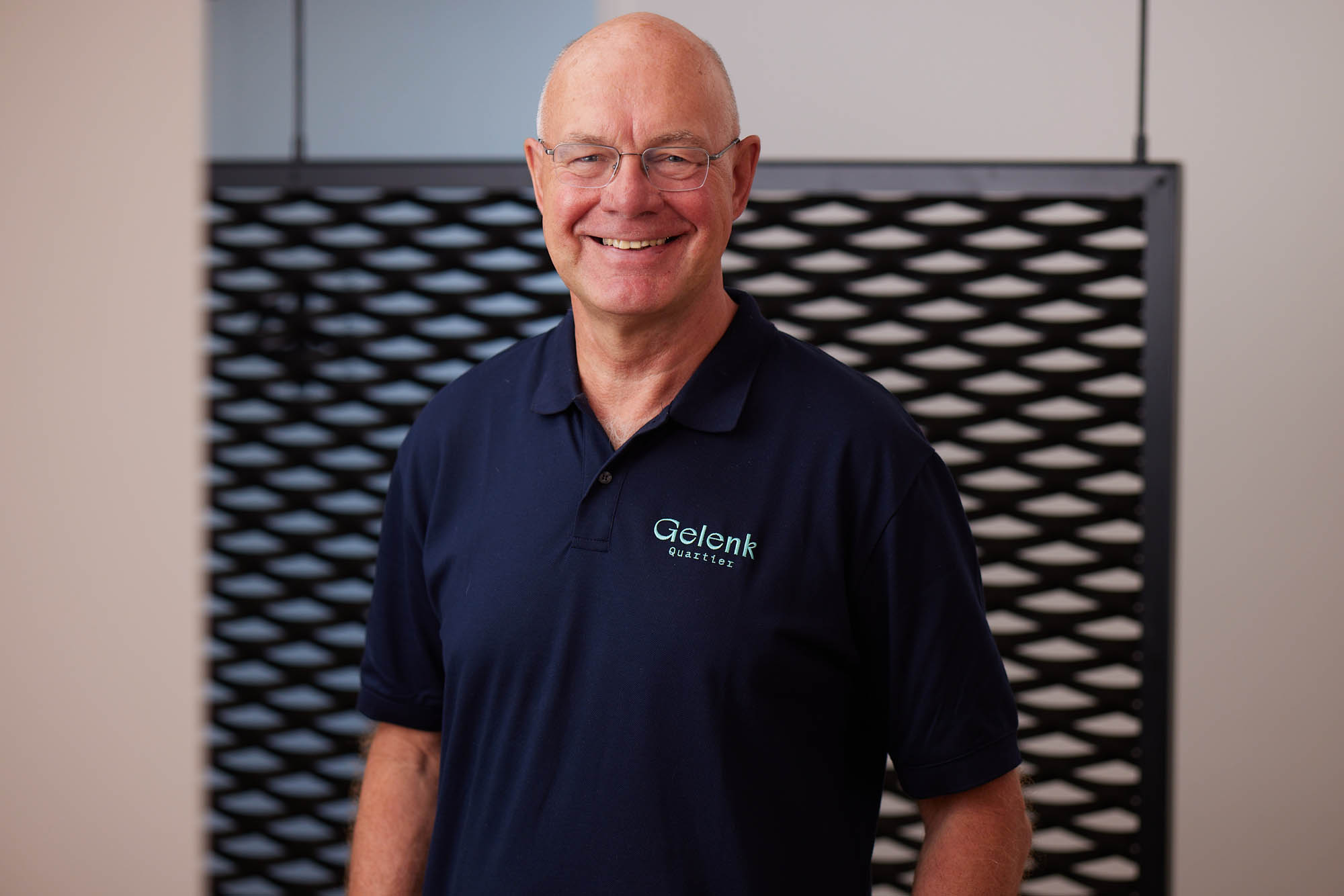 Dr. Krebs lächelt vor einer Wand stehend in die Kamera. Er trägt ein dunkelblaues Poloshirt mit dem Logo „Gelenkquartier“.