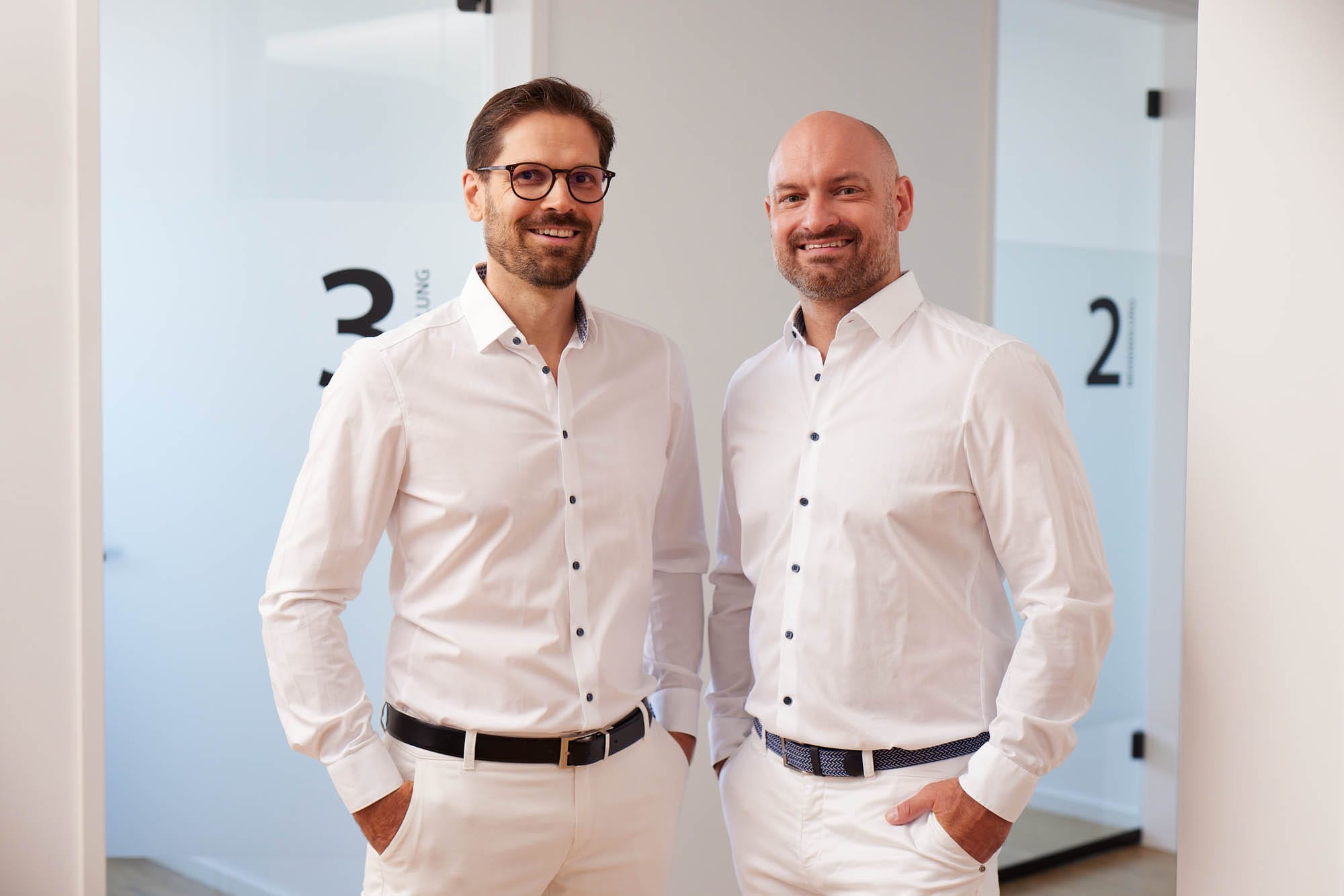 Dr. Zimmerer und Dr. Nietschke stehen in einem Flur vor Glastüren. Sie tragen helle Kleidung und lächeln in die Kamera.