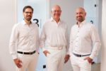 Dr. Zimmerer, Dr. Krebs und Dr. Nietschke stehen in einem Flur vor Glastüren. Sie tragen helle Kleidung und lächeln in die Kamera.