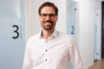 Dr. Nietschke steht in einem Flur mit Glastüren im Hintergrund. Er trägt eine Brille, ein weißes Hemd und lächelt in die Kamera.