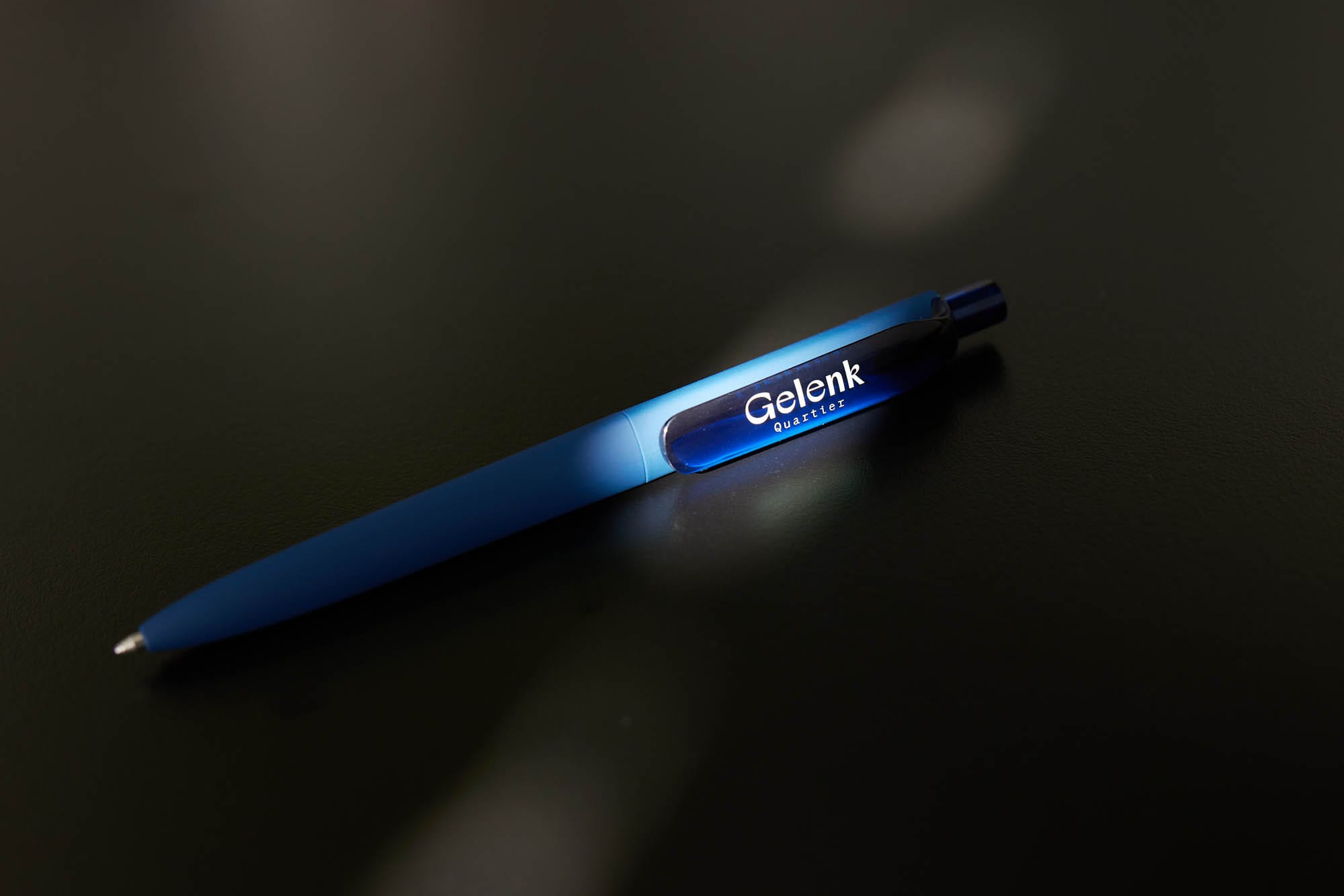 Ein blauer Kugelschreiber mit dem Logo „Gelenkquartier“ liegt auf einem dunklen Tisch. Ein Lichtstrahl hebt das Logo hervor.