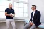 Dr. Zimmerer hält ein Modell des Beckens in der Hand. Er spricht mit einem Mann, der auf einer Untersuchungsliege sitzt.