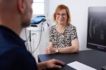 Eine Frau sitzt in einem Sprechzimmer am Tisch und spricht mit Dr. Zimmerer. Im Hintergrund steht ein medizinisches Gerät.