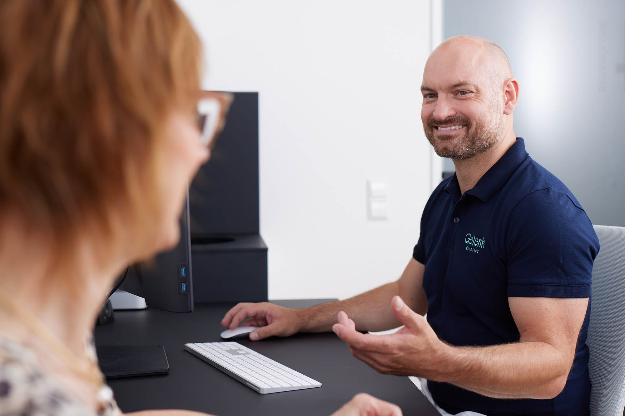 Dr. Zimmerer sitzt am Schreibtisch vor einem Computer. Er spricht lächelnd mit einer Frau, die teilweise zu sehen ist.