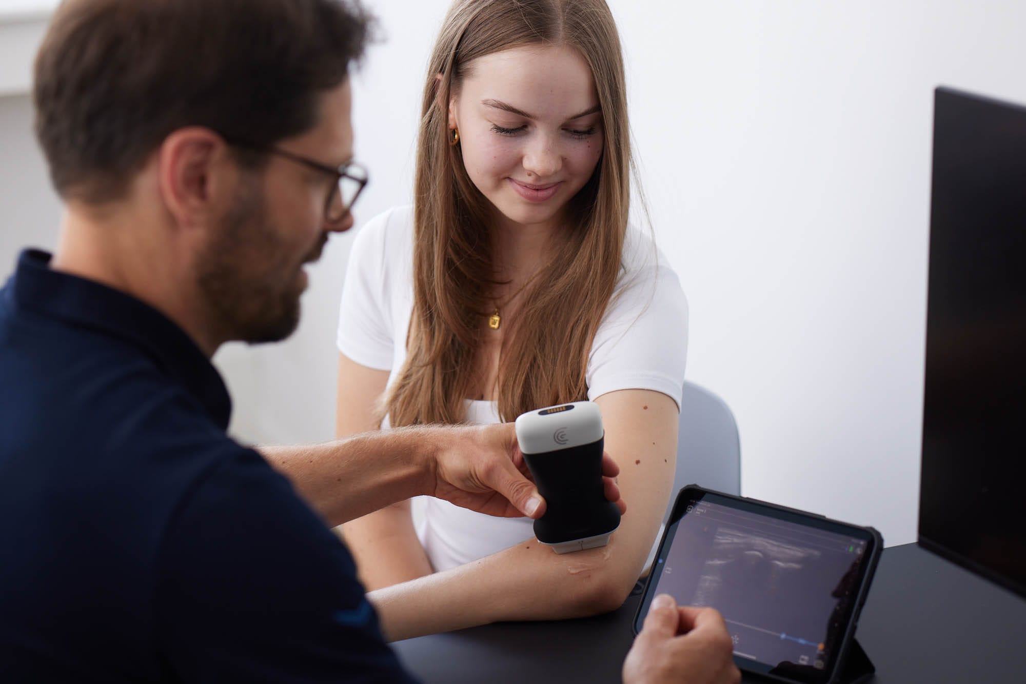 Dr. Nietschke führt bei einer Frau eine Ultraschalluntersuchung am Arm durch. Auf einem Tablet ist die Aufnahme sichtbar.