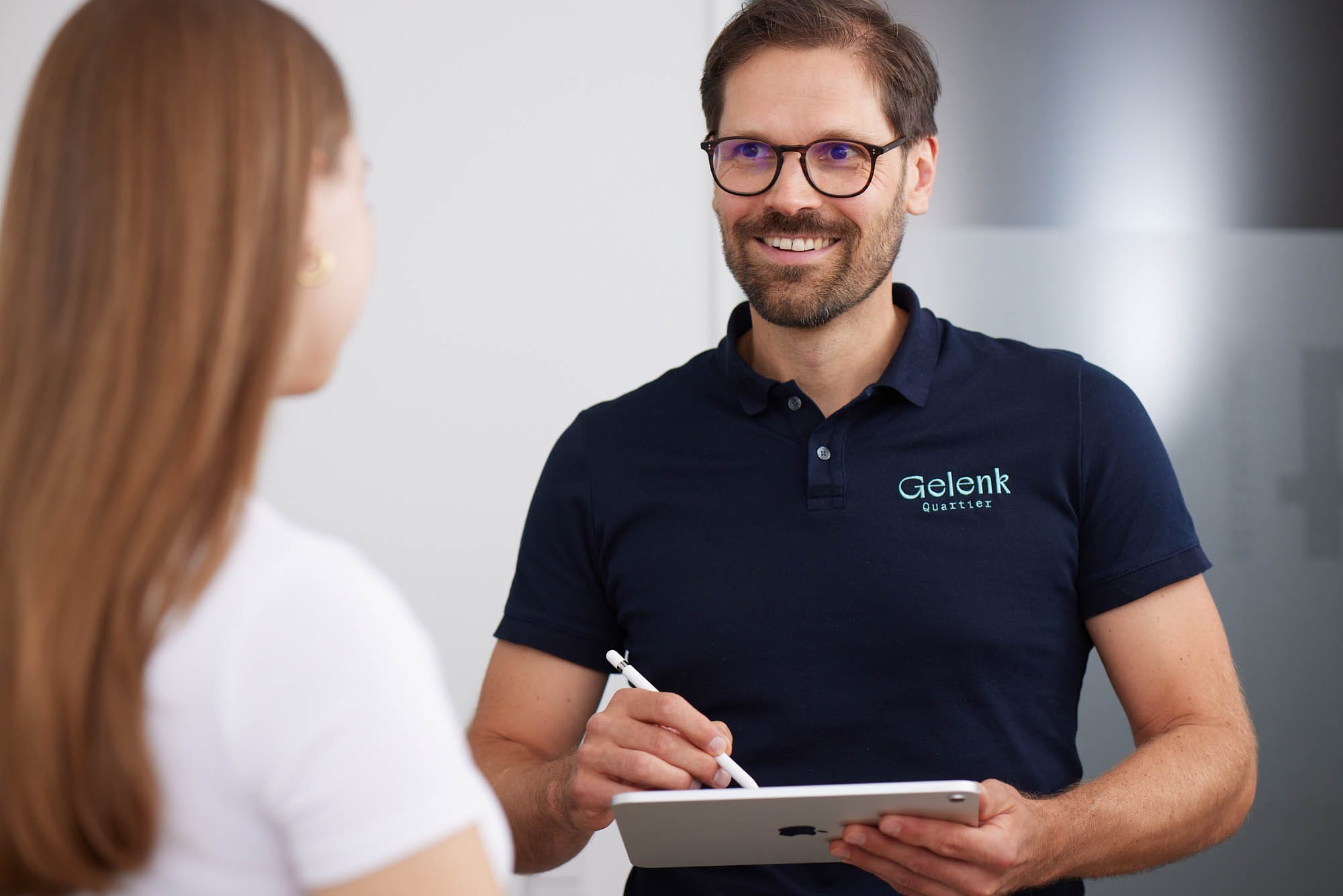 Dr. Nietschke trägt ein dunkelblaues Poloshirt und spricht mit einer Patientin während er Notizen auf dem Tablet macht.