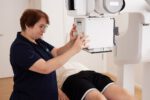 Eine medizinische Fachkraft positioniert im Vordergrund ein Röntgengerät über dem Becken eines liegenden Patienten.