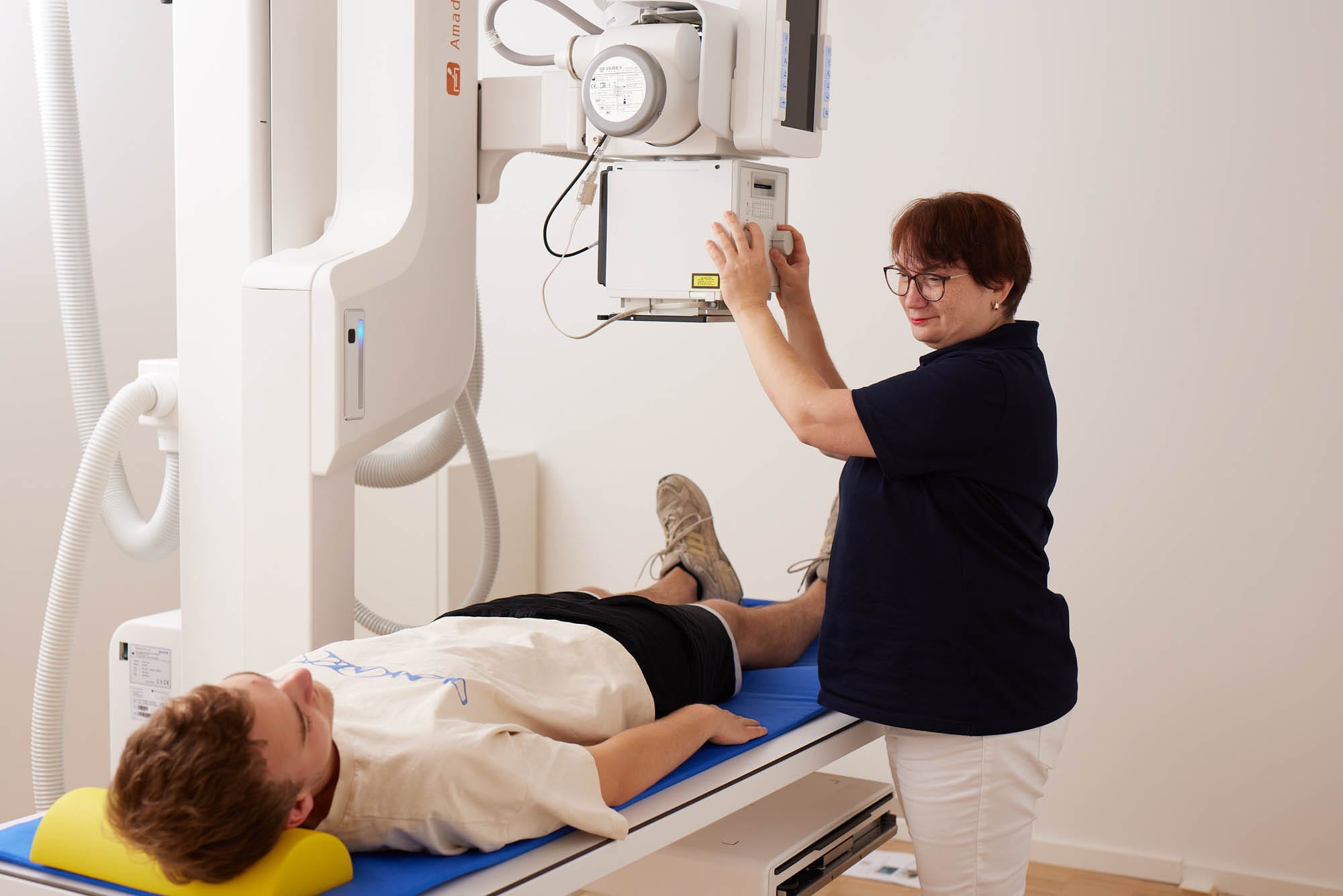Eine medizinische Fachkraft bedient ein Röntgengerät über einem Patienten, der auf einer blauen Untersuchungsliege liegt.