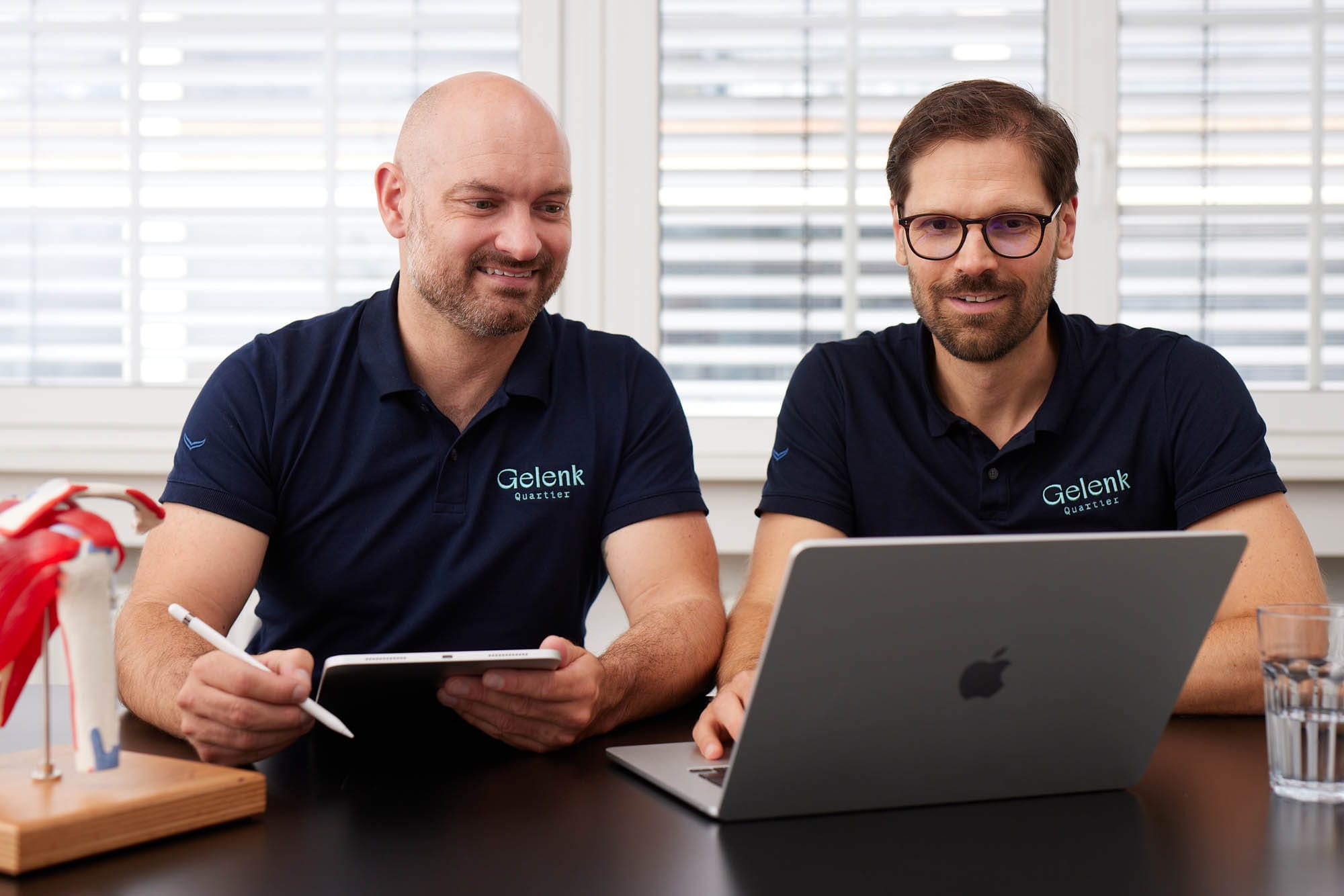 Dr. Zimmerer und Dr. Nietschke sitzen gemeinsam am Tisch. Sie besprechen etwas an einem Laptop und Tablet.